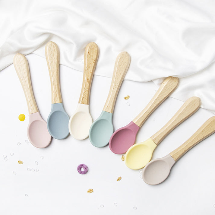 A row of five colorful silicone spoons with smooth wooden handles, resting on a white tablecloth.