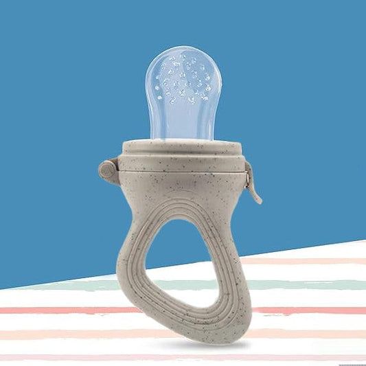 A silicone baby food feeder with a gray handle and a see-trough mesh pouch sits on a white table. The feeder has a spoon-like shape, with the pouch at the wide end and the handle at the narrow end. The pouch is empty.